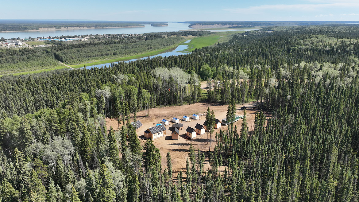An aerial shot of a clear in a forest at the edge of a river. The clearing shows a large cabin, gazebo, five cabins, five tents, a portable, and solar panels.