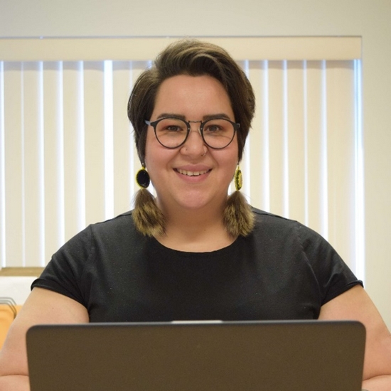 Alexia souriant devant un ordinateur portable. Elle a des cheveux courts et foncés, des lunettes et des boucles d'oreilles ornées de fourrure.
