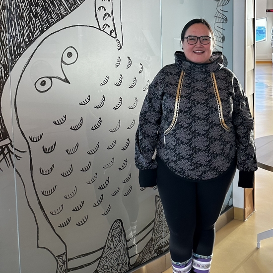 Kamele devant une œuvre d'art à la Station canadienne de recherche dans l'Extrême-Arctique, portant un kalikuk (haut de type coquille souple) et des kamiit (bottes) brodées représentatives de la région de Kitikmeot.